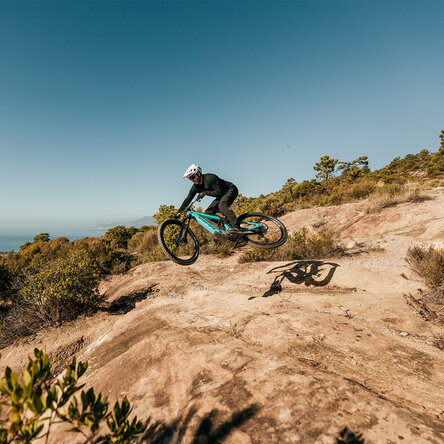 Un mountain biker su una mountain bike elettronica blu-verde salta su un trail terroso, su un pendio soleggiato con vista mare sullo sfondo.