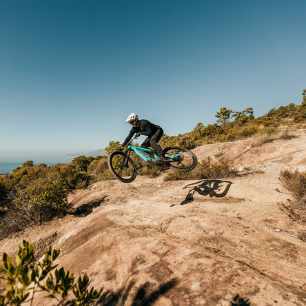 Ein Mountainbiker auf einem blau-grünen E-Mountainbike springt über einen erdigen Trail an einem sonnigen Berghang mit Meerblick im Hintergrund.