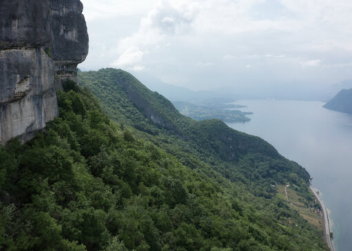 Vue sur un paysage de montagne boisé, dominé par des falaises abruptes et un grand lac en arrière-plan sous un ciel nuageux.