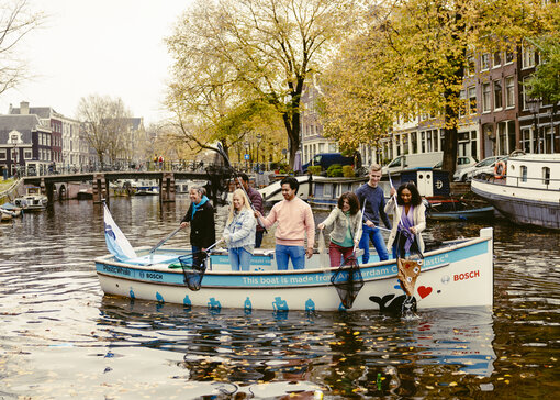 Meerdere mensen zitten op een bootje op een gracht en vissen met schepnetten dingen uit het water.