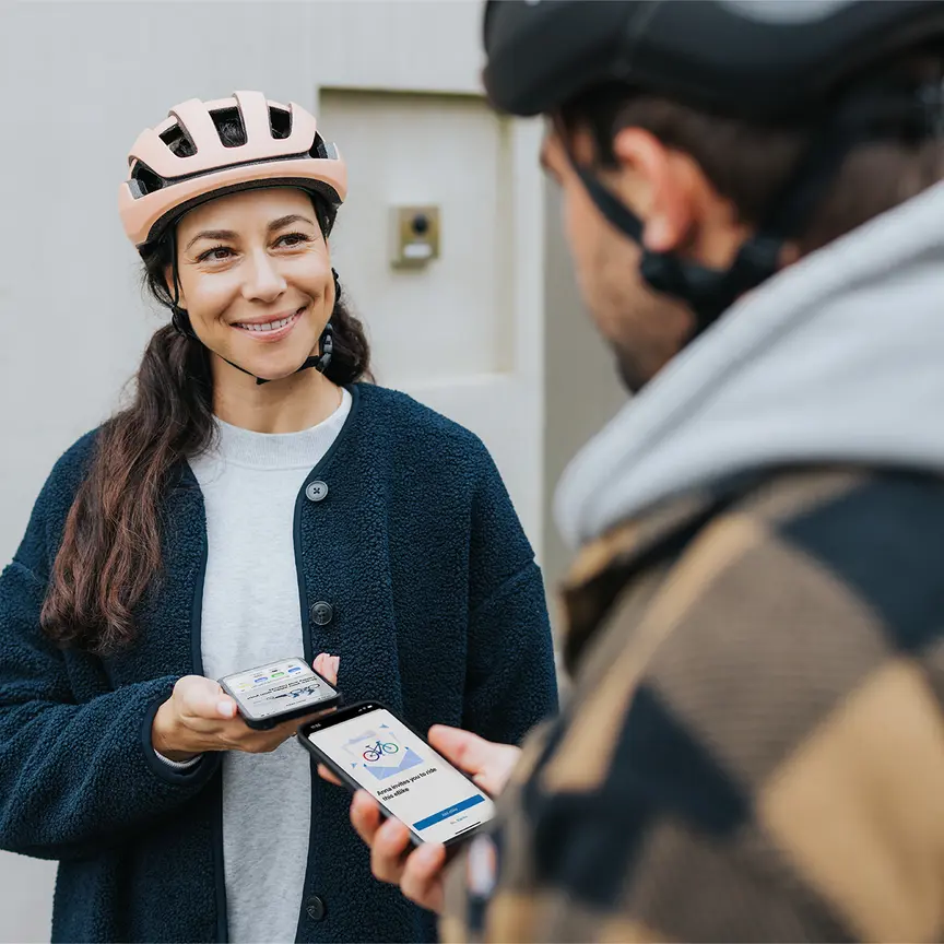 Une femme et un homme se font face en souriant et connectent leurs smartphones dans l’application eBike Flow. Ils portent tous les deux un casque de vélo.