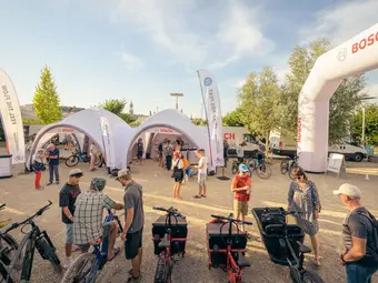 Some people are seen from a wide-angle perspective at a cycling festival. In the background, Bosch tents and flags are visible.