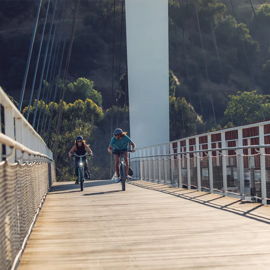 Two people ride e-bikes across a bridge with railings and wire ropes.