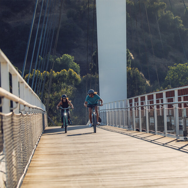 Two people ride e-bikes across a bridge with railings and wire ropes.