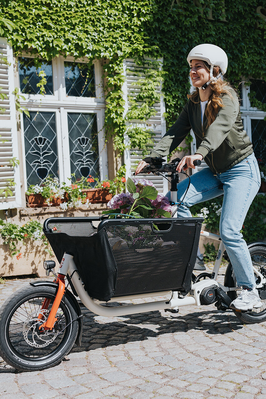 Eine Person fährt ein Lasten-E-Bike und trägt einen weißen Helm sowie eine grüne Jacke und blaue Jeans. Im Korb des Fahrrads befinden sich Blumen. Im Hintergrund ist ein Gebäude mit weißen Fensterläden und grüner Efeubewachsung zu sehen.