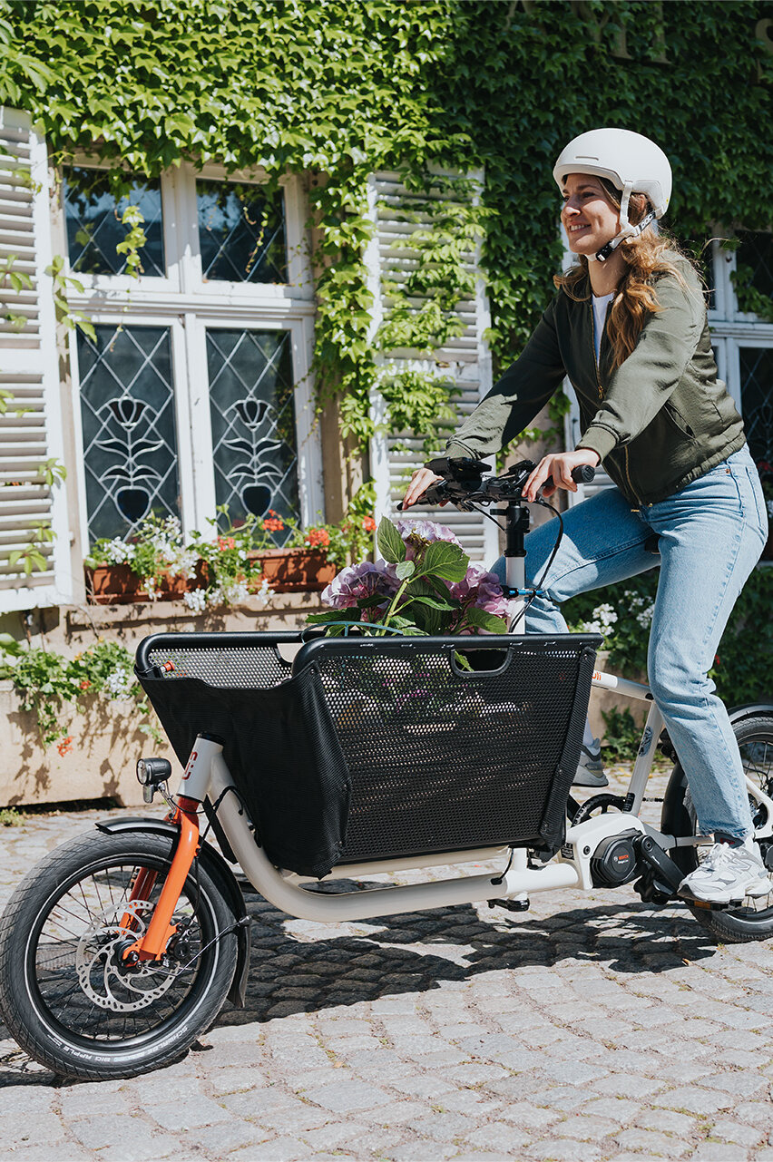 Eine Person fährt ein Lasten-E-Bike und trägt einen weißen Helm sowie eine grüne Jacke und blaue Jeans. Im Korb des Fahrrads befinden sich Blumen. Im Hintergrund ist ein Gebäude mit weißen Fensterläden und grüner Efeubewachsung zu sehen.