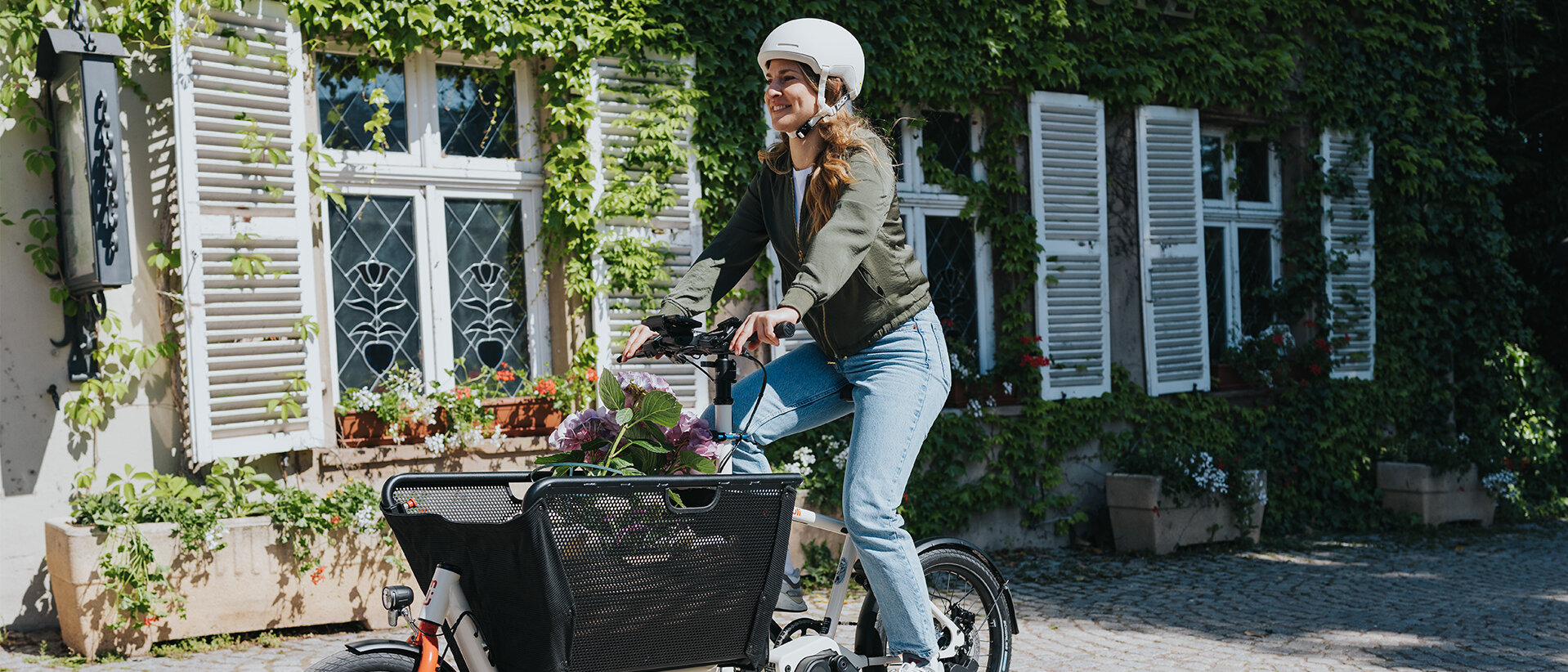 Eine Person fährt ein Lasten-E-Bike und trägt einen weißen Helm sowie eine grüne Jacke und blaue Jeans. Im Korb des Fahrrads befinden sich Blumen. Im Hintergrund ist ein Gebäude mit weißen Fensterläden und grüner Efeubewachsung zu sehen.