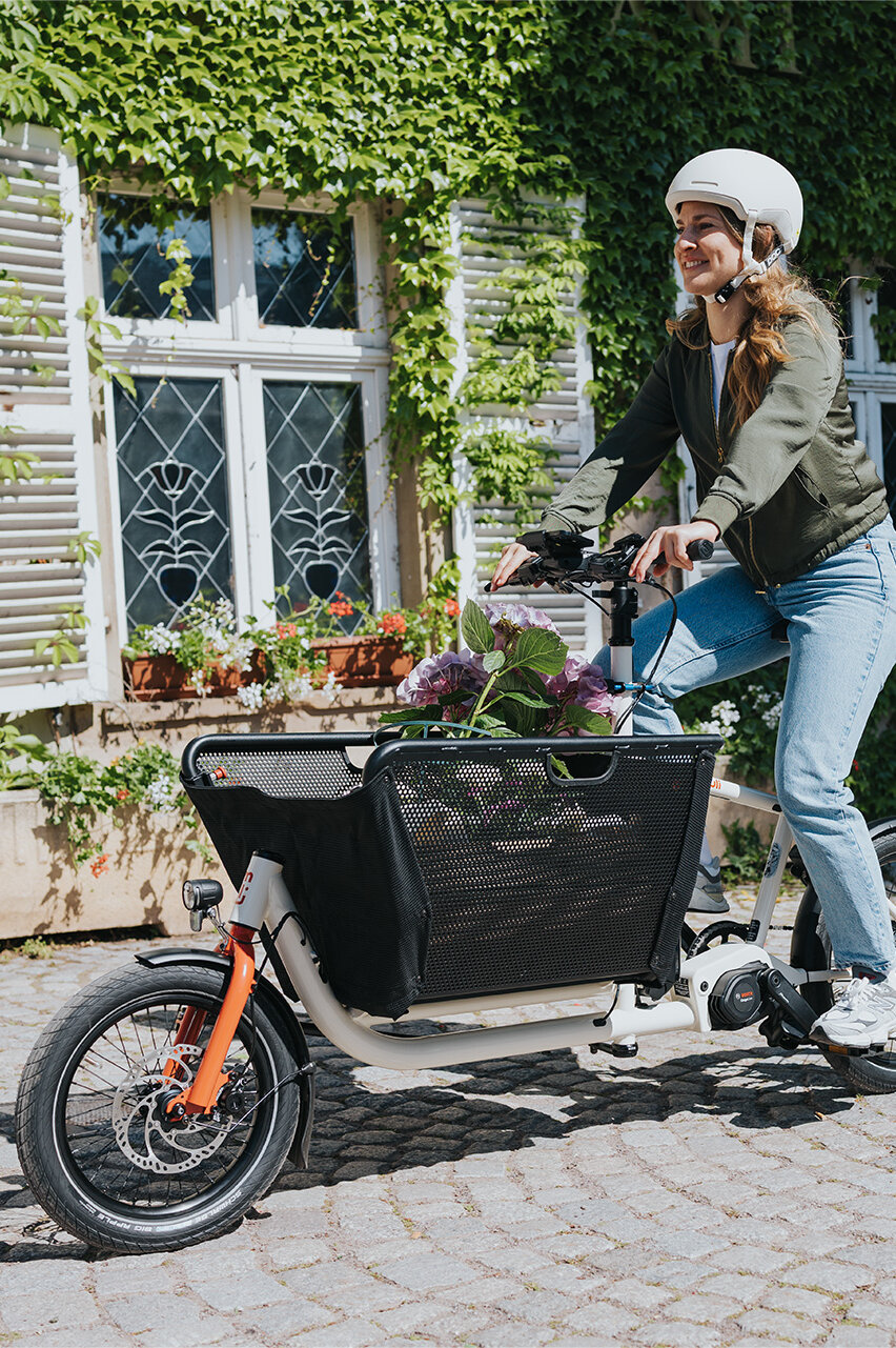 Eine Person fährt ein Lasten-E-Bike und trägt einen weißen Helm sowie eine grüne Jacke und blaue Jeans. Im Korb des Fahrrads befinden sich Blumen. Im Hintergrund ist ein Gebäude mit weißen Fensterläden und grüner Efeubewachsung zu sehen.