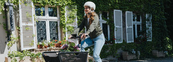Eine Person fährt ein Lasten-E-Bike und trägt einen weißen Helm sowie eine grüne Jacke und blaue Jeans. Im Korb des Fahrrads befinden sich Blumen. Im Hintergrund ist ein Gebäude mit weißen Fensterläden und grüner Efeubewachsung zu sehen.