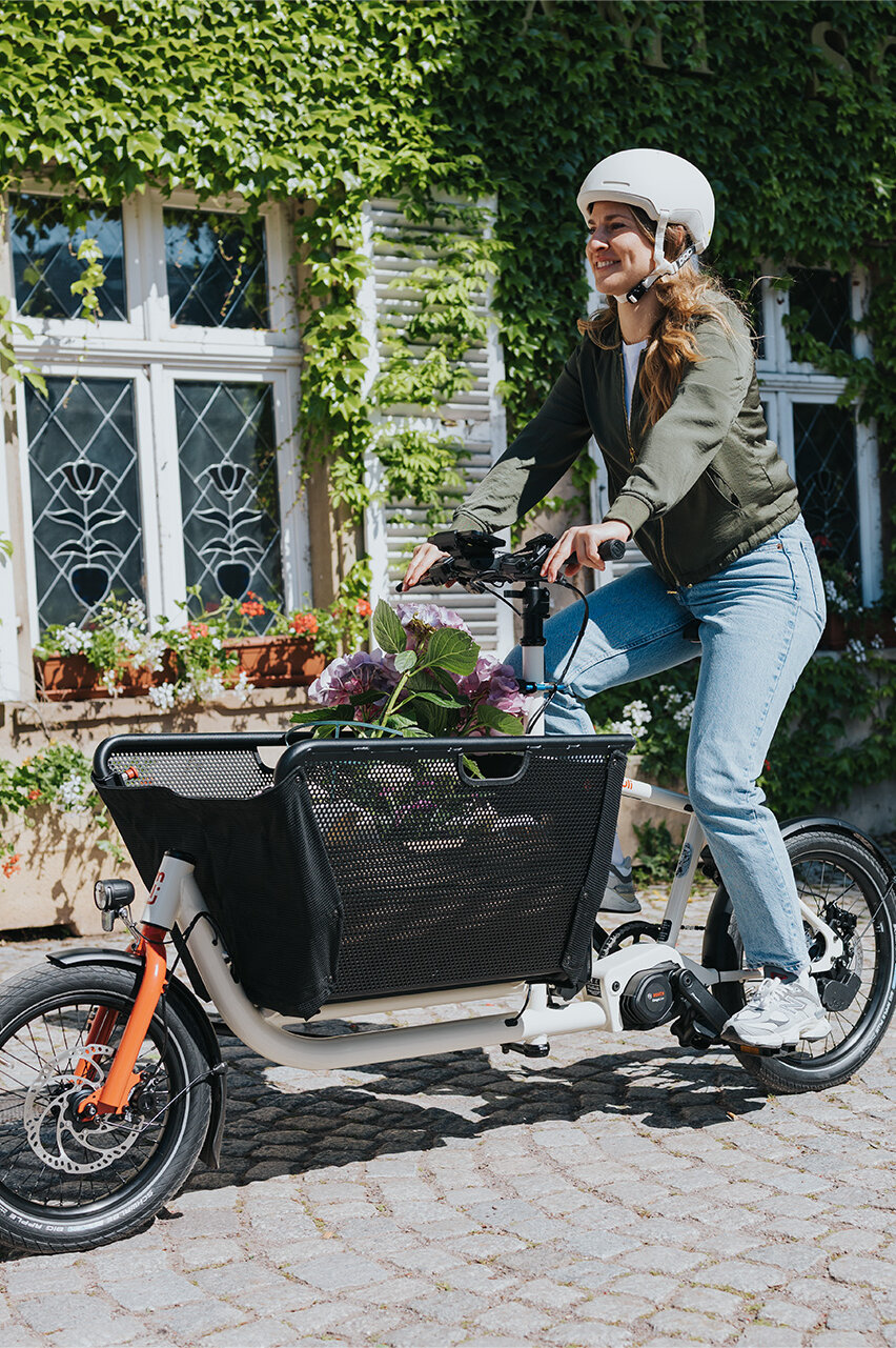 Eine Person fährt ein Lasten-E-Bike und trägt einen weißen Helm sowie eine grüne Jacke und blaue Jeans. Im Korb des Fahrrads befinden sich Blumen. Im Hintergrund ist ein Gebäude mit weißen Fensterläden und grüner Efeubewachsung zu sehen.