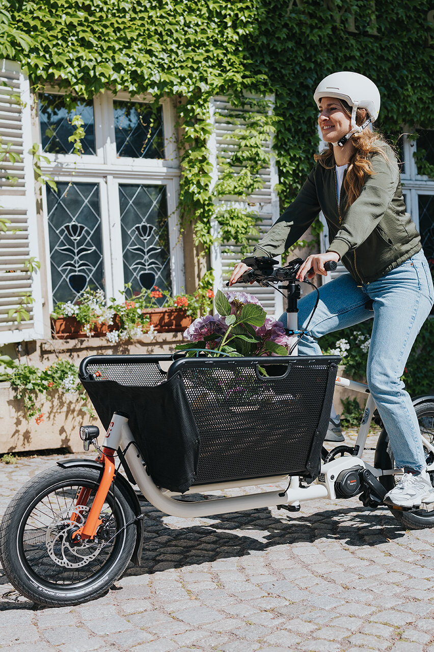 A person is riding a cargo e-bike, wearing a white helmet, a green jacket, and blue jeans. Flowers fill the basket of the bicycle. In the background, you see a building with white shutters and green ivy.