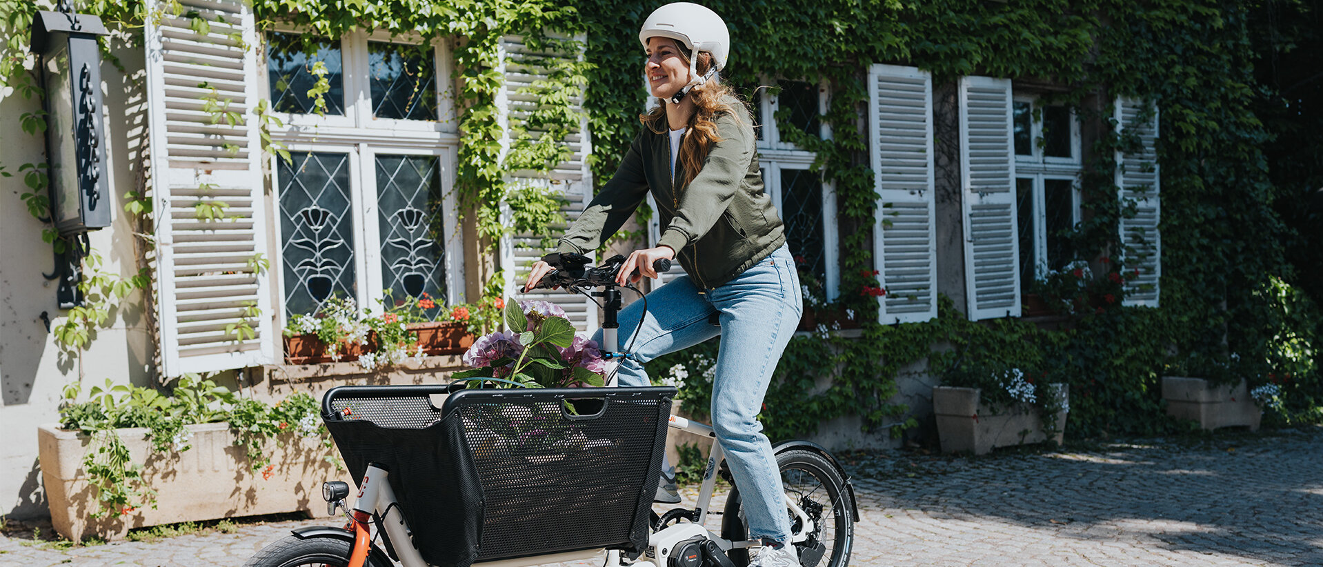 Eine Person fährt ein Lasten-E-Bike und trägt einen weißen Helm sowie eine grüne Jacke und blaue Jeans. Im Korb des Fahrrads befinden sich Blumen. Im Hintergrund ist ein Gebäude mit weißen Fensterläden und grüner Efeubewachsung zu sehen.