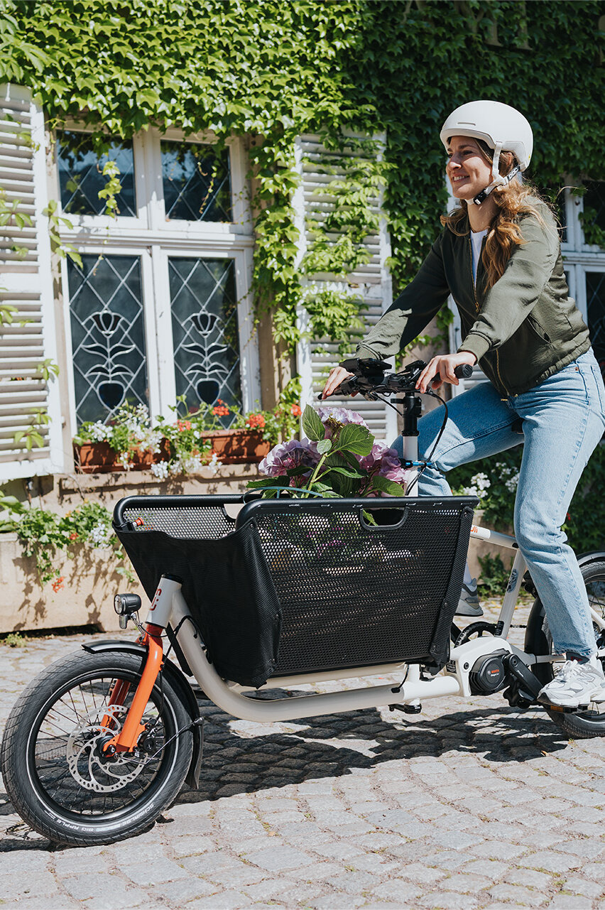 Eine Person fährt ein Lasten-E-Bike und trägt einen weißen Helm sowie eine grüne Jacke und blaue Jeans. Im Korb des Fahrrads befinden sich Blumen. Im Hintergrund ist ein Gebäude mit weißen Fensterläden und grüner Efeubewachsung zu sehen.