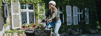 Eine Person fährt ein Lasten-E-Bike und trägt einen weißen Helm sowie eine grüne Jacke und blaue Jeans. Im Korb des Fahrrads befinden sich Blumen. Im Hintergrund ist ein Gebäude mit weißen Fensterläden und grüner Efeubewachsung zu sehen.