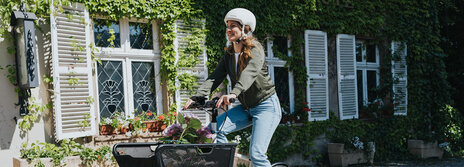 Eine Person fährt ein Lasten-E-Bike und trägt einen weißen Helm sowie eine grüne Jacke und blaue Jeans. Im Korb des Fahrrads befinden sich Blumen. Im Hintergrund ist ein Gebäude mit weißen Fensterläden und grüner Efeubewachsung zu sehen.