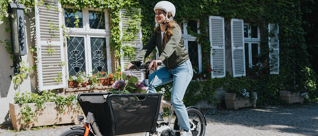 Eine Person fährt ein Lasten-E-Bike und trägt einen weißen Helm sowie eine grüne Jacke und blaue Jeans. Im Korb des Fahrrads befinden sich Blumen. Im Hintergrund ist ein Gebäude mit weißen Fensterläden und grüner Efeubewachsung zu sehen.