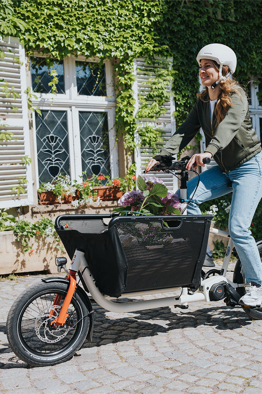 Eine Person fährt ein Lasten-E-Bike und trägt einen weißen Helm sowie eine grüne Jacke und blaue Jeans. Im Korb des Fahrrads befinden sich Blumen. Im Hintergrund ist ein Gebäude mit weißen Fensterläden und grüner Efeubewachsung zu sehen.