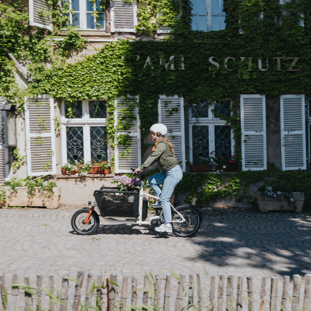 Une femme avec un casque roule sur un VAE Cargo le long d’une maison couverte de lierre.