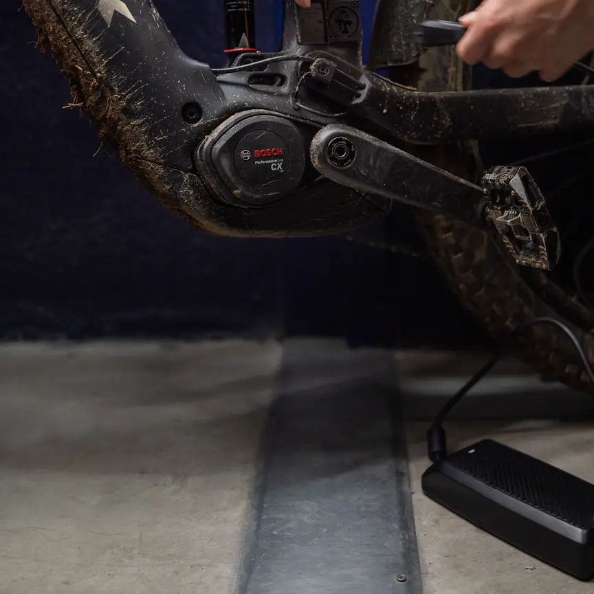 Nærbillede, der viser et mudderplettet eMountainbike, som bliver tilsluttet til opladning. En hånd fører stikket fra 12A-opladeren ind i ladeporten på rammen, direkte over Performance Line CX-motoren fra Bosch.