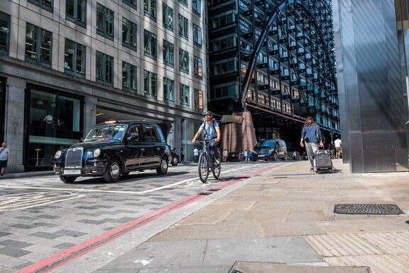 Mann mit eBike-Helm fährt auf Pedelec durch die Londoner City an einem parkenden Taxi vorbei