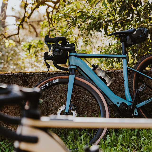 Two eBikers sit on a wall, smiling as they look at a smartphone together. Next to them leans a Gravel eBike.