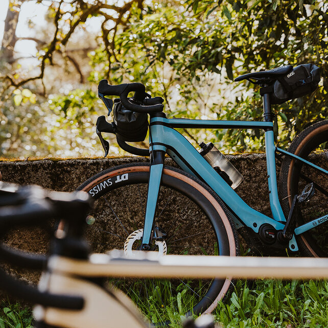 Two eBikers sit on a wall, smiling as they look at a smartphone together. Next to them leans a Gravel eBike.