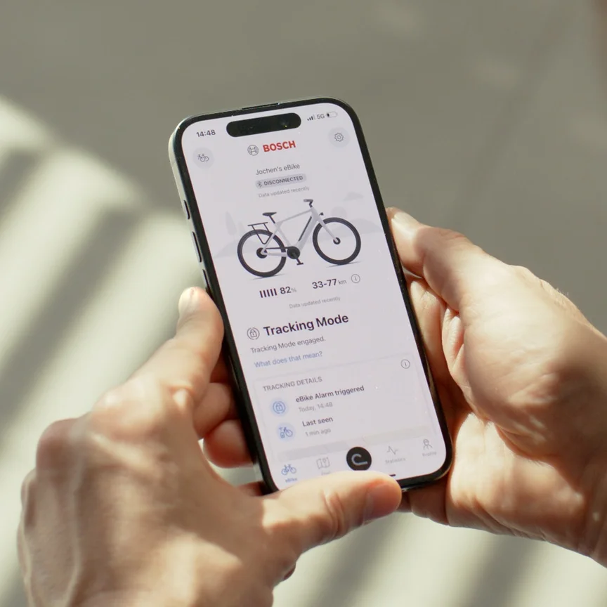 Osoba trzyma smartfon z otwartą aplikacją eBike Flow Bosch. Ekran wyświetlacza pokazuje aktywowany tryb Tracking oraz uruchomiony alarm rower ze wspomaganiem elektrycznym / e-rower, który jest funkcją ochrony rower ze wspomaganiem elektrycznym / e-rower przed kradzieżą.