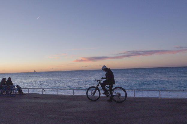 Un cycliste VAE fait une pause et regarde son smartphone fixé au guidon. À l’arrière-plan, on voit la mer au coucher du soleil ainsi que des personnes assises sur des bancs.