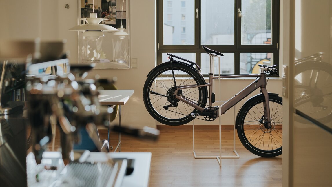 A modern eBike with a hub motor is displayed on a minimalist stand in a stylish, bright room with a wooden floor and a large window. The shot is taken from the perspective of a kitchen, with a professional espresso machine in the blurred foreground, giving the scene a lifestyle vibe.