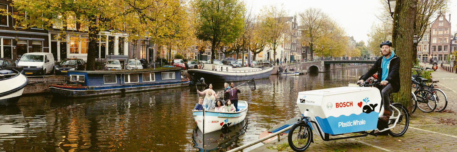 Een man zit aan de oever van een kanaal op een Cargo Bike met het opschrift "Bosch Plastic Whale". In de gracht staan enkele mensen op een kleine boot en kijken in de camera, waarbij het Bosch-logo op de boot te zien is.