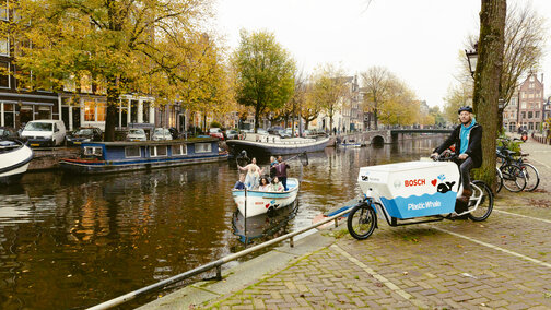 Een man zit aan de oever van een kanaal op een Cargo Bike met het opschrift "Bosch Plastic Whale". In de gracht staan enkele mensen op een kleine boot en kijken in de camera, waarbij het Bosch-logo op de boot te zien is.