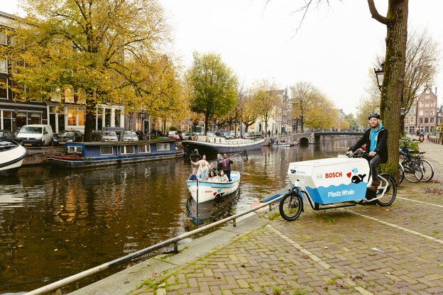Een man zit aan de oever van een kanaal op een Cargo Bike met het opschrift "Bosch Plastic Whale". In de gracht staan enkele mensen op een kleine boot en kijken in de camera, waarbij het Bosch-logo op de boot te zien is.