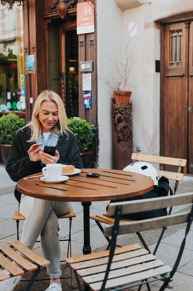En kvinna sitter vid ett bord på ett kafé och tittar på sin smartphone. I bakgrunden står hennes vita eBike.