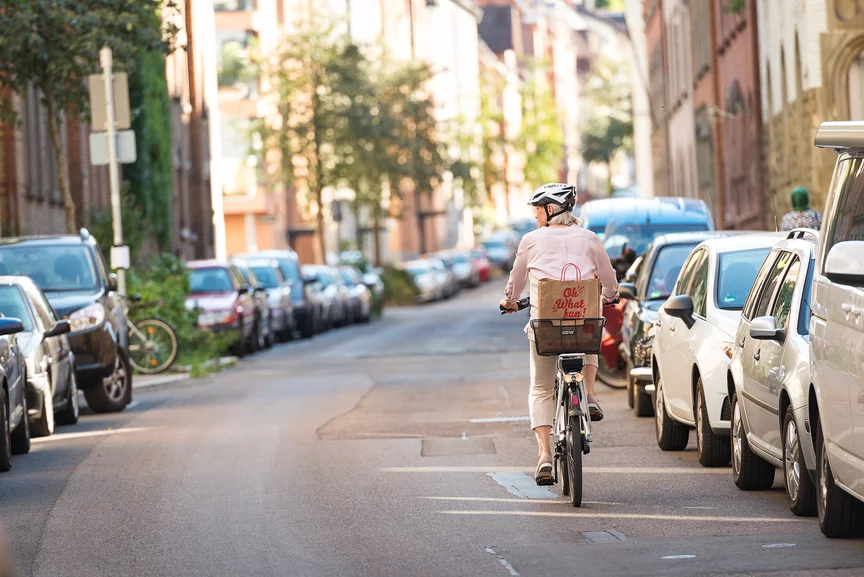 Eine eBike Fahrerin fährt mit ihrem eBike neben parkenden Autos eine Straße hinunter. Auf ihrem Gepäckträger hat sie einen Korb, in welchem eine Tüte mit der Aufschrift "Oh What fun" steht.