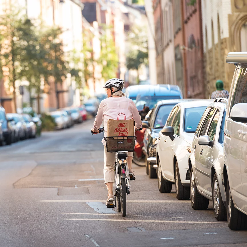 Rowerzystka na rowerze ze wspomaganiem elektrycznym / e-rower jedzie ulicą obok zaparkowanych samochodów. Na bagażniku ma koszyk, w którym znajduje się torba z napisem "Oh What fun".