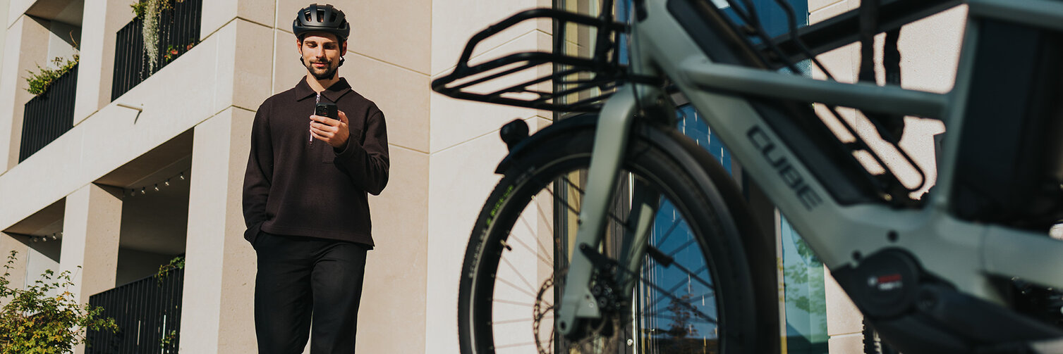 Een man, helemaal in het zwart gekleed, loopt glimlachend naar een eBike terwijl hij op zijn smartphone kijkt.