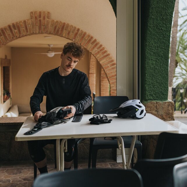 Ein Mountainbiker sitzt an einem Tisch auf einer überdachten Terrasse und packt seinen 12A Charger in seine Tasche. Sein Helm und seine Handschuhe liegen bereit. Im Hintergrund sind gemauerte Bögen und Palmen zu sehen, was eine mediterrane Atmosphäre vermittelt.