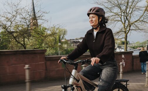 En smilende kvinne sykler elsykkel på en rød sykkelsti langs en urban havnefront. Bildet formidler gleden ved å sykle i byen til hverdags på en solrik dag.
