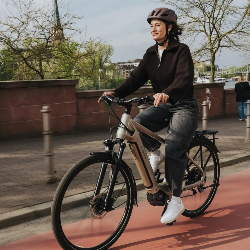 Usměvavá žena jede na svém eBike po červené cyklostezce podél městského nábřeží. Záběr vyjadřuje radost z každodenní jízdy ve městě za slunečného dne.
