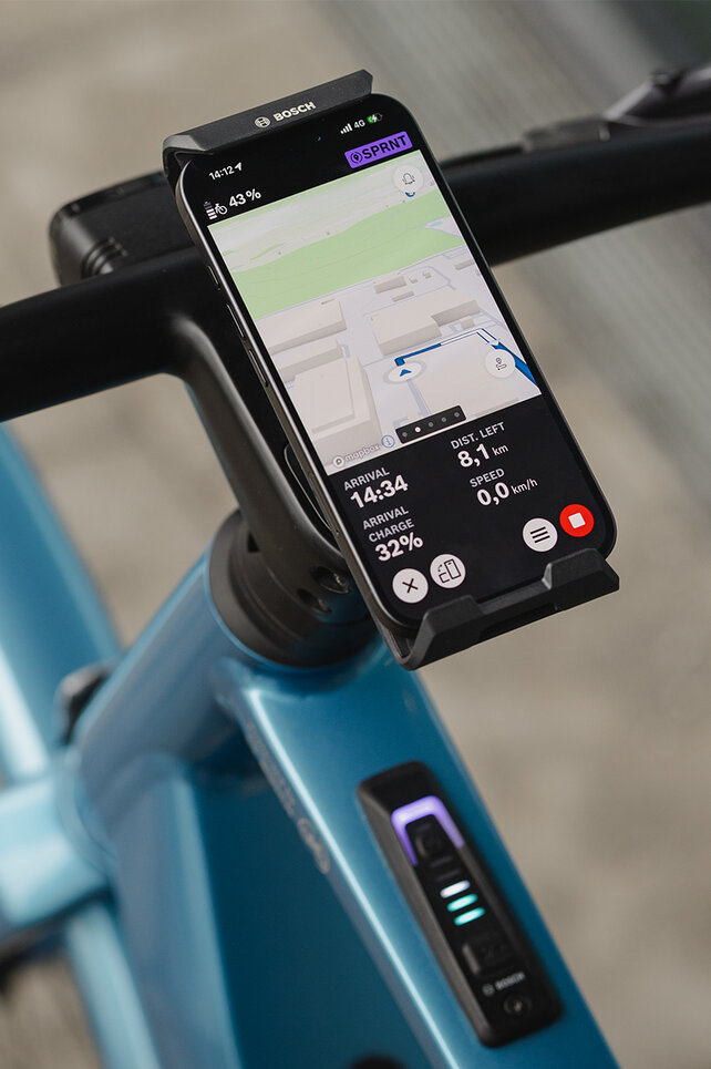 Primer plano de un SmartphoneGrip vertical fijado al manillar. El smartphone está en modo navegación y muestra una guía de ruta.