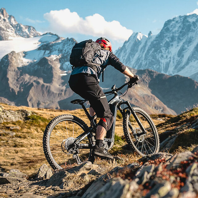 Un conducteur VTT à assistance électrique de l'arrière avec un sac à dos sur son VTT à assistance électrique roule sur des rochers, des montagnes peuvent être vues en arrière-plan.