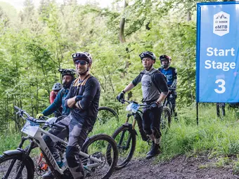 Several eBiker are at the starting point of an eMTB race with their eBikes. A green forest is visible in the background.