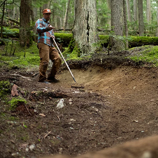 Scott Veach se tient sur un sol terreux au milieu de la forêt et prépare le terrain avec un râteau.