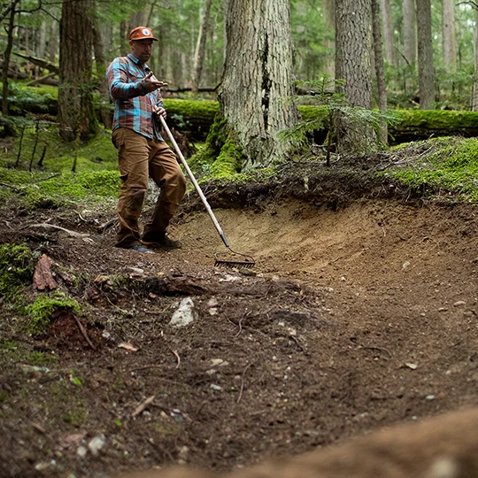 Scott Veach stands on earthy ground in the middle of the forest, using a rake to prepare the soil.