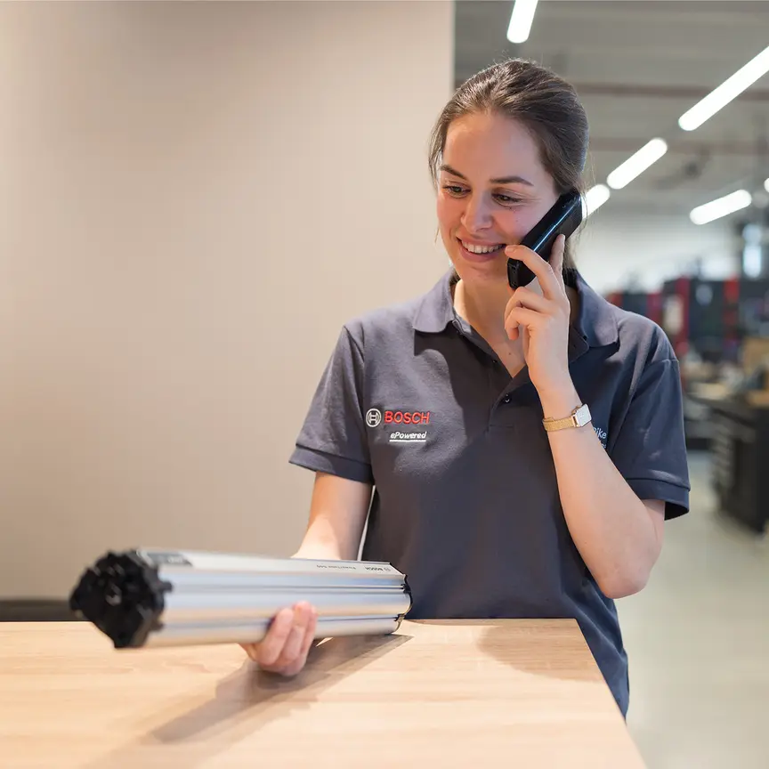 En kvinne med en Bosch-skjorte holder en PowerTube i hånden. Samtidig snakker hun i telefonen. I bakgrunnen ser du et verkstedmiljø.