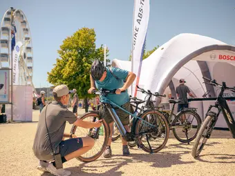 A Bosch specialist and a man are inspecting the front wheel suspension of an eBike. In the background, there is a Bosch tent, a Ferris wheel, and a tree.