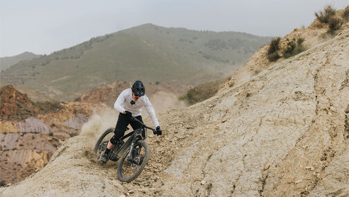 An eMountain bike rider descends a steep rocky slope, kicking up dust. Loose stones are visible along the path, and a mountain rises in the background.