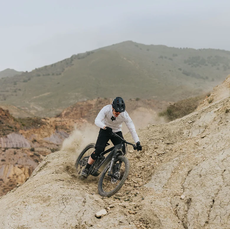 An eMountain bike rider descends a steep rocky slope, kicking up dust. Loose stones are visible along the path, and a mountain rises in the background.