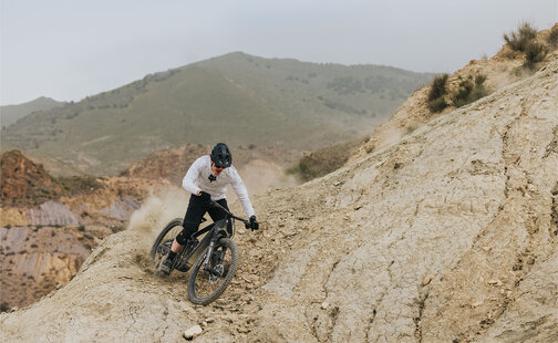 An eMountain bike rider descends a steep rocky slope, kicking up dust. Loose stones are visible along the path, and a mountain rises in the background.