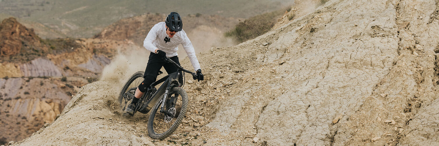 Ein eMountainbike Fahrer fährt einen steilen Steinabhang hinab und wirbelt dabei Staub auf. Auf dem Weg sind lose Steine zu erkennen, im Hintergrund erkennt man einen Berg.